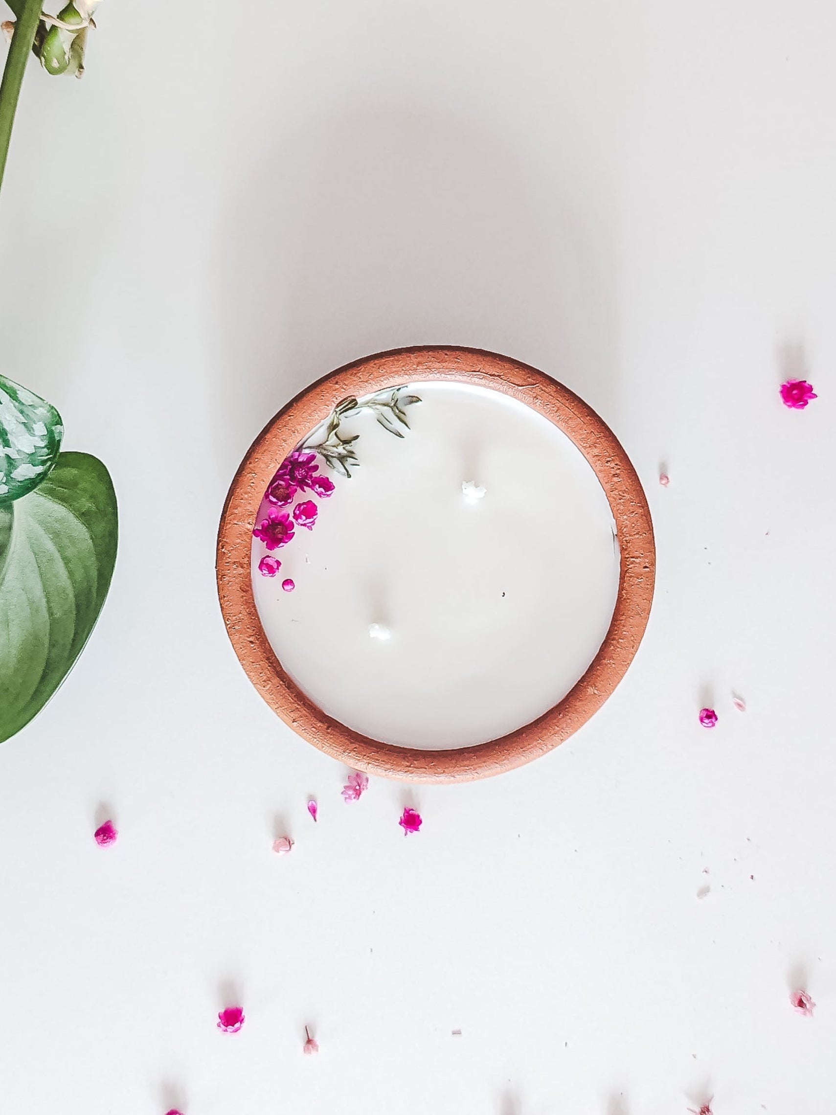 A top-down view of a white soy wax candle in a round wooden bowl with a small pink floral accent, set on a white surface with scattered pink petals and a green plant stem on the left.