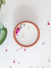 A top-down view of a white soy wax candle in a round wooden bowl with a small pink floral accent, set on a white surface with scattered pink petals and a green plant stem on the left.